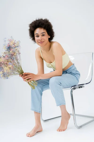 Barefoot woman in pants and top holding flowers while sitting on chair on grey background