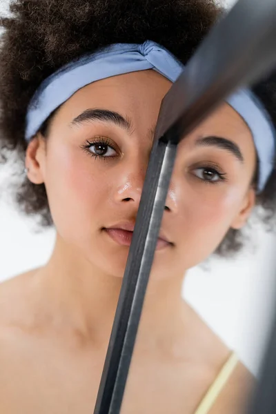 African american woman with headband looking at camera near mirror isolated on grey