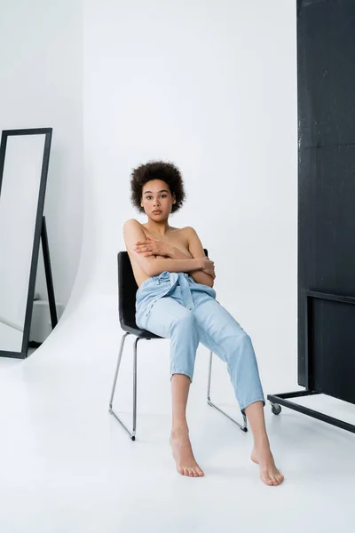 Shirtless african american woman in pants sitting on chair on grey background