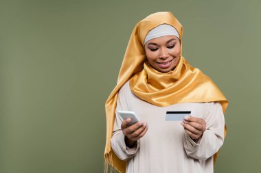 Smiling multiracial woman in hijab using smartphone and credit card isolated on green  clipart