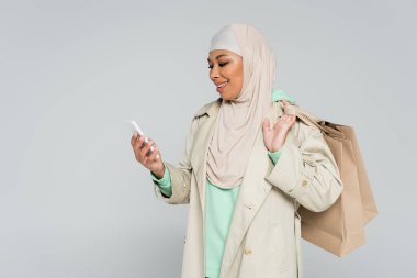 Trençkotlu, tesettürlü, tesettürlü, alışveriş çantalı ve gri renkli akıllı telefona bakan çok kültürlü bir kadın.