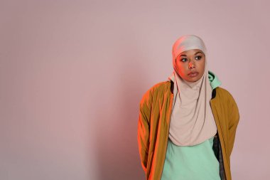 thoughtful multiracial woman in yellow jacket and muslim hijab looking away on pinkish grey background clipart