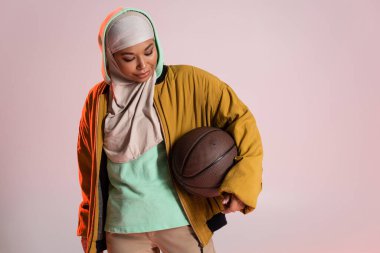 fashionable multiracial woman in hijab and yellow bomber jacket holding basketball isolated on pinkish grey clipart