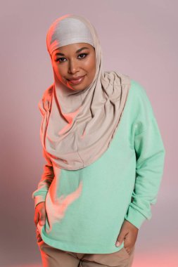 fashionable multiracial woman in hijab and green long sleeve shirt smiling at camera on pinkish grey background clipart