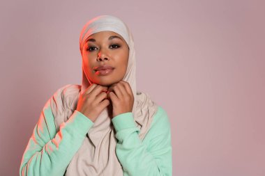 positive multiracial woman in green long sleeve shirt adjusting traditional hijab isolated on pinkish grey clipart