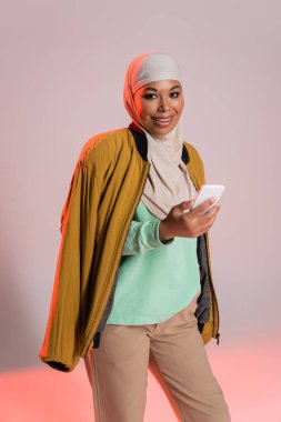 Genç ve çok ırklı kadın tesettürlü ve sarı bombacı ceketli gri ve pembe arka planda akıllı telefon tutan.