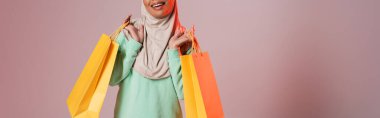 cropped view of happy multiracial muslim woman holding yellow shopping bags on pinkish grey background, banner clipart