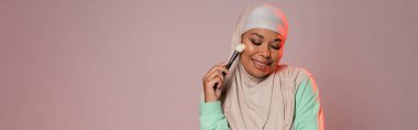smiling multiracial woman in hijab holding cosmetic brush while standing with closed eyes isolated on pinkish grey, banner clipart