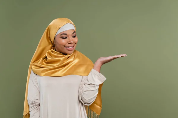 Positive multiracial woman in hijab pointing with hand isolated on green