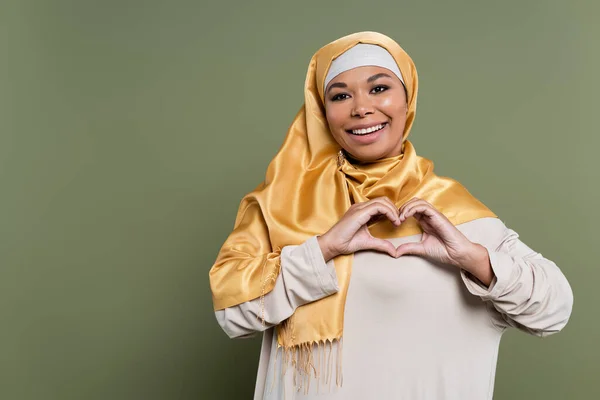Smiling multiracial woman in hijab showing heart sign on green background