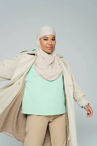 穿着穆斯林头巾和战壕外套的无忧无虑的多种族女人对着用灰色隔离的相机摆姿势微笑 — 图库照片