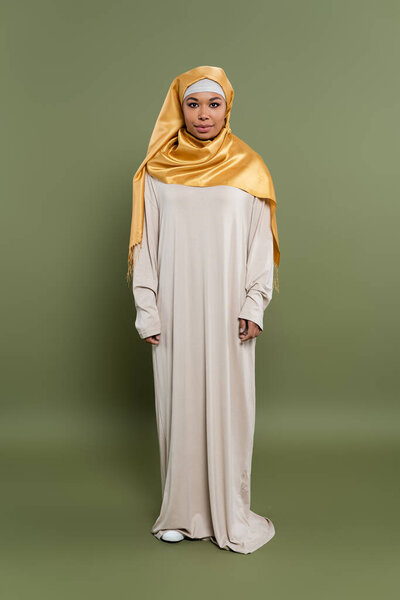 full length of multiracial muslim woman in yellow silk hijab and abaya dress standing on olive green background