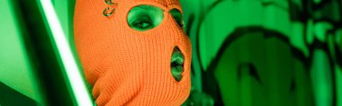 Parlak neon lambanın yanında örülmüş turuncu kar maskeli seksi bir kadın portresi ve grafitiyle yeşil bir duvar.