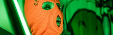 portrait of sexy woman in knitted orange balaclava near bright neon lamp and green wall with graffiti, banner clipart