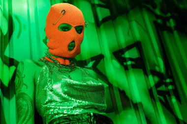 low angle view of young woman in balaclava and sexy top looking at camera in green lighting near wall with graffiti clipart
