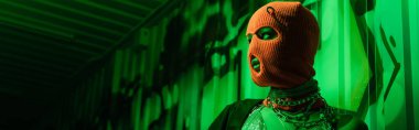 sexy anonymous woman in orange balaclava and silver necklaces looking away in green light near wall with graffiti, banner clipart