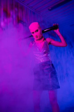 provocative woman in balaclava and black leather skirt standing with baseball bat in purple lighting with smoke clipart