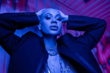 Baştan çıkarıcı kısa saçlı, siyah ceketli, gümüş boyunlu, kafaya dokunan ve mavi ışıklandırmalı kameraya bakan kadının düşük açılı görüntüsü.