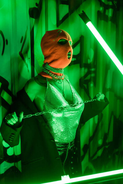татуированная женщина в блестящем топе и вязаной балаклаве позирует с металлической цепью возле зеленой стены с граффити и неоновой лампой