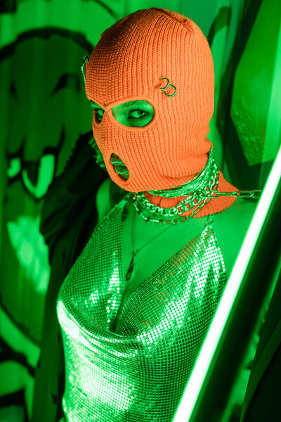 sexy woman in knitted balaclava and shiny top posing near neon lamp and wall with graffiti in green lighting
