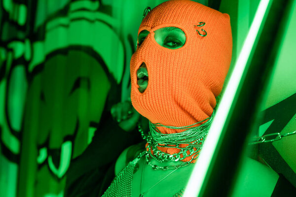 passionate woman in orange balaclava and silver neck chains looking at camera near vibrant neon lamp and wall with graffiti
