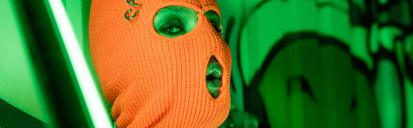 portrait of sexy woman in knitted orange balaclava near bright neon lamp and green wall with graffiti, banner