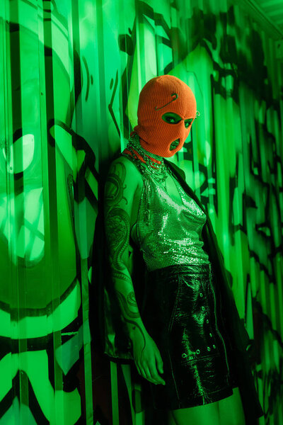 sexy woman in black leather skirt and silver top with orange balaclava standing near green wall with graffiti
