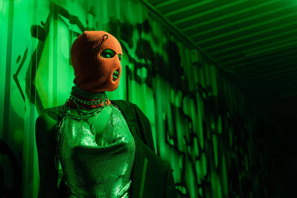 анонимная сексуальная женщина в оранжевой балаклаве и серебряном топе, смотрящая на стену с граффити в зеленом свете