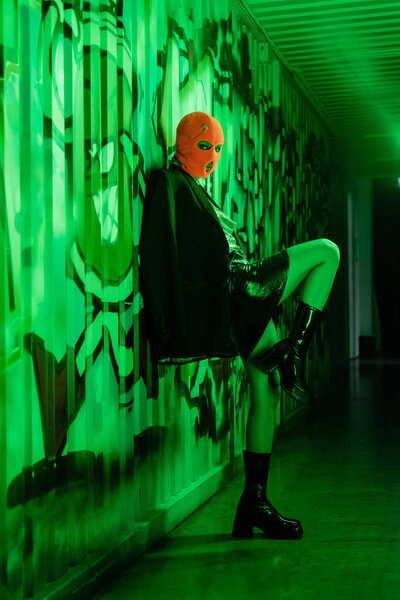 full length of sexy woman in stylish leather boots and orange balaclava standing near wall with graffiti in green lighting