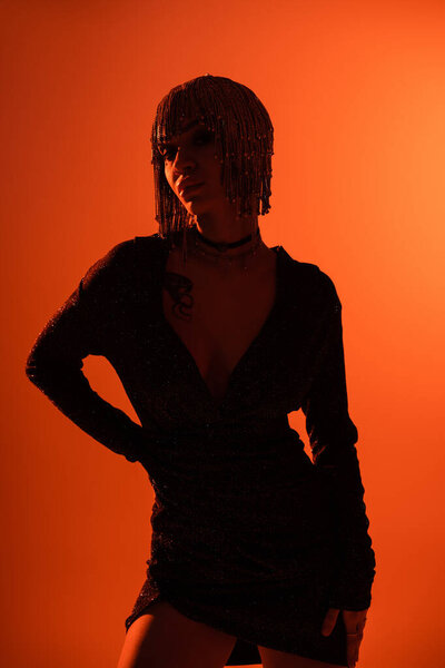 татуированная женщина в черном платье и металлическом головном уборе позирует с рукой на оранжевом фоне