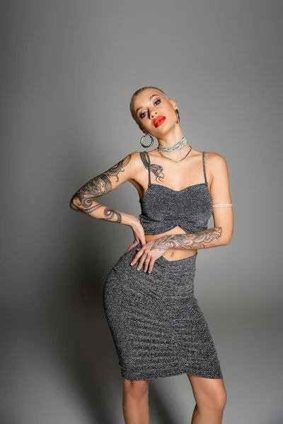 tattooed woman with red lips wearing lurex skirt with crop top and posing with hand on hip on grey background