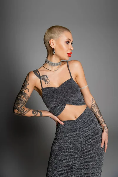 seductive woman in lurex crop top and necklaces looking away while posing with hand on hip on grey background