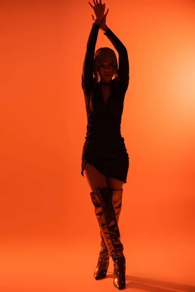 stock image full length of slender woman in black sexy dress and long boots posing with arms up on orange background