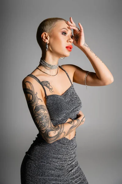 tattooed woman with short hair and red lips holding hand near face and posing in glamour clothes isolated on grey