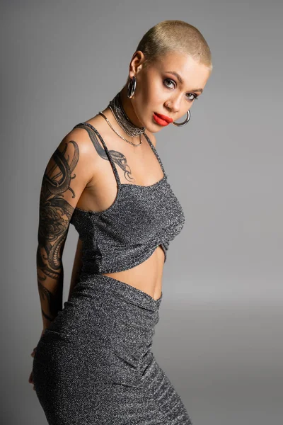 stock image sexy tattooed woman with short hair and red lips wearing lurex skirt and crop top while looking at camera isolated on grey