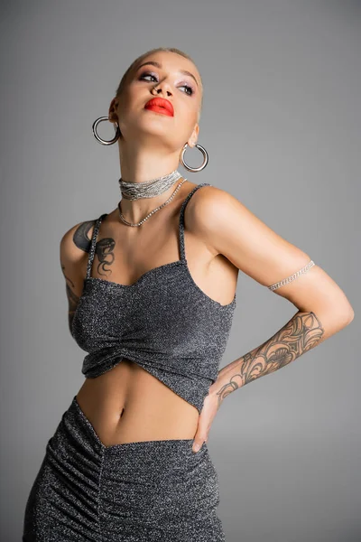 extravagant tattooed woman with hoop earrings and red lips posing with hand on waist and looking away isolated on grey