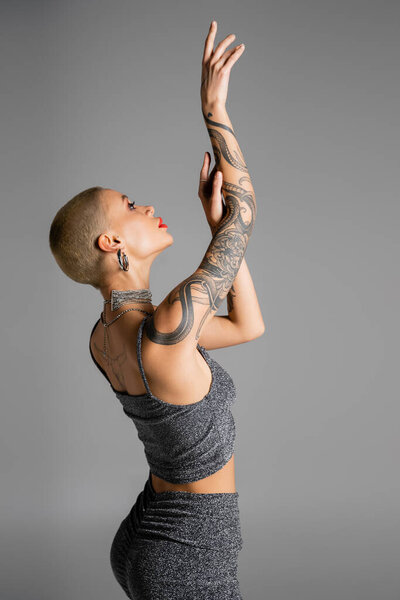 side view of trendy woman in lurex crop top posing with raised tattooed hands isolated on grey