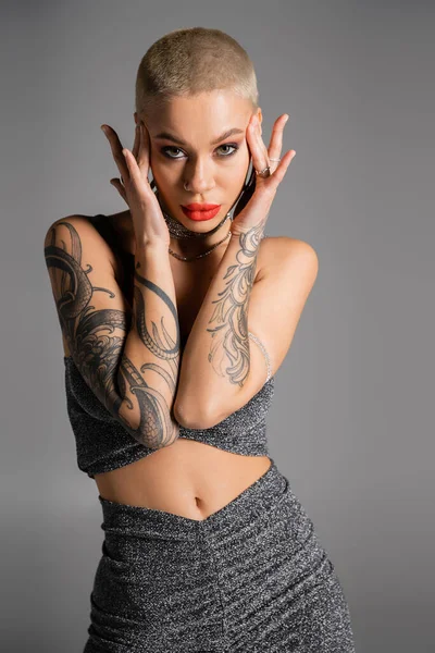 seductive tattooed woman with short hair and bright red lips touching head and looking at camera isolated on grey