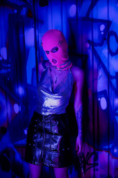 passionate woman in balaclava and silver top looking at camera near wall with graffiti in blue light