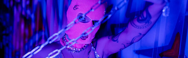 сексуальная татуированная женщина в балаклаве держа серебряные цепи возле стены с граффити в синем и фиолетовом свете, баннер