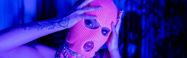 young tattooed woman touching balaclava and looking at camera near graffiti in blue neon light, banner