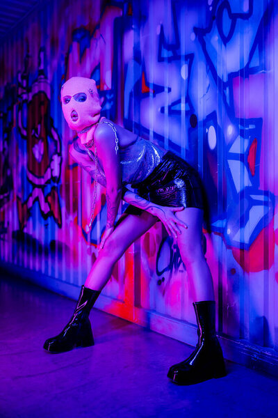 seductive woman in balaclava and black leather boots posing near colorful graffiti in blue and purple neon light