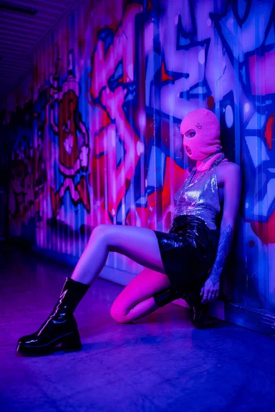 バラクラバでセクシーな女性の完全な長さと流行の服は青と紫のネオンライトでカラフルな落書きと壁の近くに座って — ストック写真