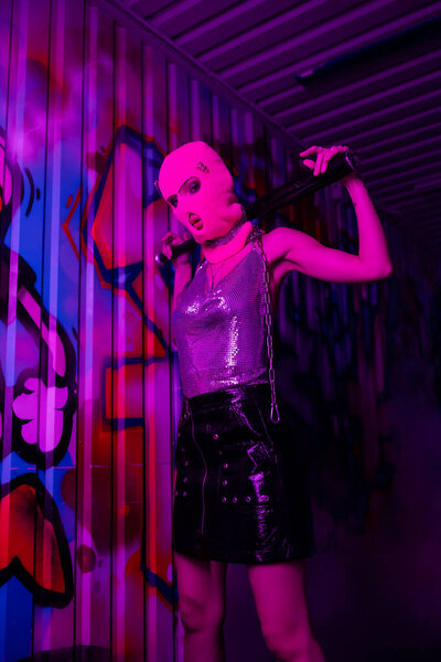 stylish woman in balaclava and metallic top posing with baseball bat near wall with graffiti in purple neon light