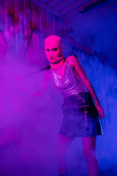 穿着粉色巴拉克拉瓦和金属上衣的迷人女人 站在墙上涂鸦附近 拿着紫色烟雾弥漫的棒球棒 — 图库照片