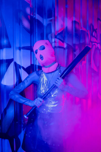 страстная женщина в балаклаве стоящая с бейсбольной битой возле голубой стены с граффити в фиолетовом дыму