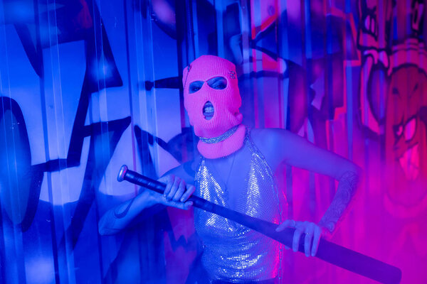 провокационная женщина в балаклаве и серебристом топе гримасит бейсбольной битой рядом с граффити в синем и фиолетовом свете