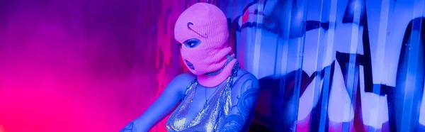 anonymous tattooed woman in balaclava near wall with graffiti in blue and pink lighting with smoke, banner