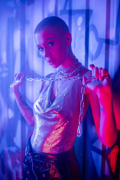 молодая и сексуальная женщина в блестящей верхней позе с серебряной цепью возле стены с граффити в синем туманном свете 