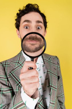 Wide angle view of stylish man in jacket holding magnifying glass isolated on yellow   clipart
